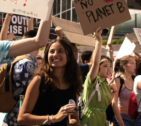 A group of young people march outdoors at a climate protest, holding signs with messages like NO PLANET B. One woman in front smiles, and the crowd appears energetic and engaged.